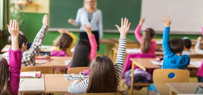  Жвава класна кімната з дітьми, які піднімають руки, і вчителем, що стоїть біля дошки, символізуючи енергію та натхнення для креативного привітання з днем народження директора школи.
